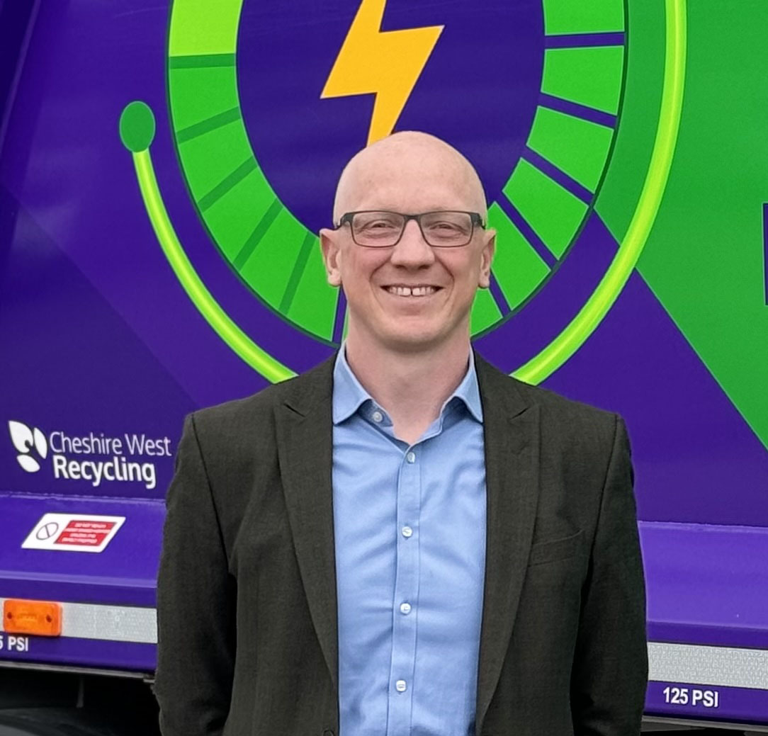 A man with glasses, blue shirt and black suit jacket stands in front of a purple refuse truck with green icon.